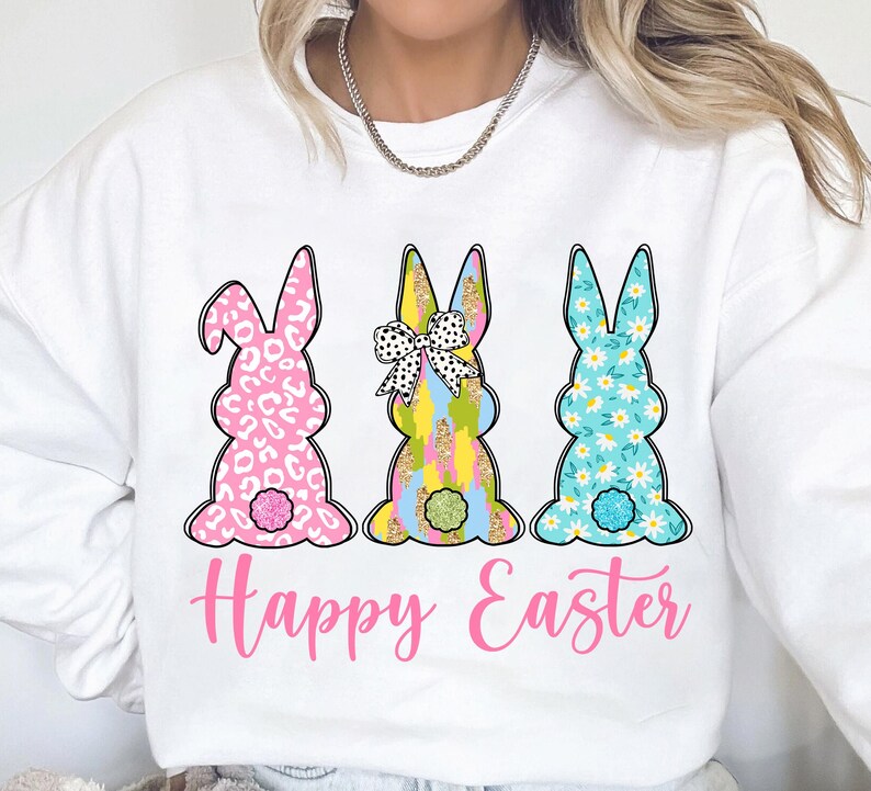 May include: White sweatshirt with a pink, blue, and yellow bunny rabbit design. The bunnies are wearing bows and have different patterns. The text "Happy Easter" is printed in pink below the bunnies.