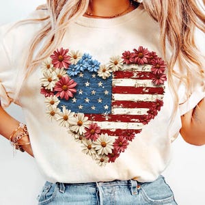 May include: Cream-colored sweatshirt featuring a heart-shaped design of the American flag, adorned with red, white, and blue flowers. The flag design has a distressed, vintage look, with visible stars and stripes.