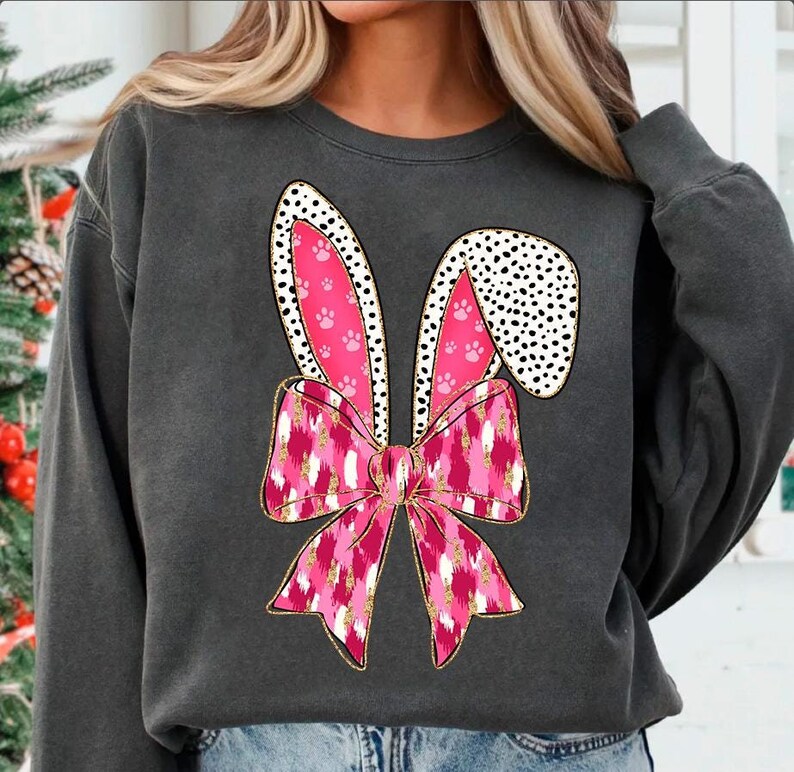 May include: A charcoal grey sweatshirt featuring a pink and white bunny ear design. The ears have a pink paw print pattern and the bow is pink with a white and gold glitter pattern.