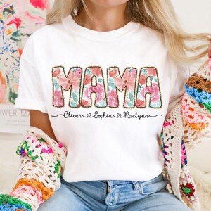 May include: White t-shirt with the word "MAMA" in pink and green floral letters. The names "Oliver", "Sophia", and "Raelynn" are written below the word "MAMA" with hearts between each name.