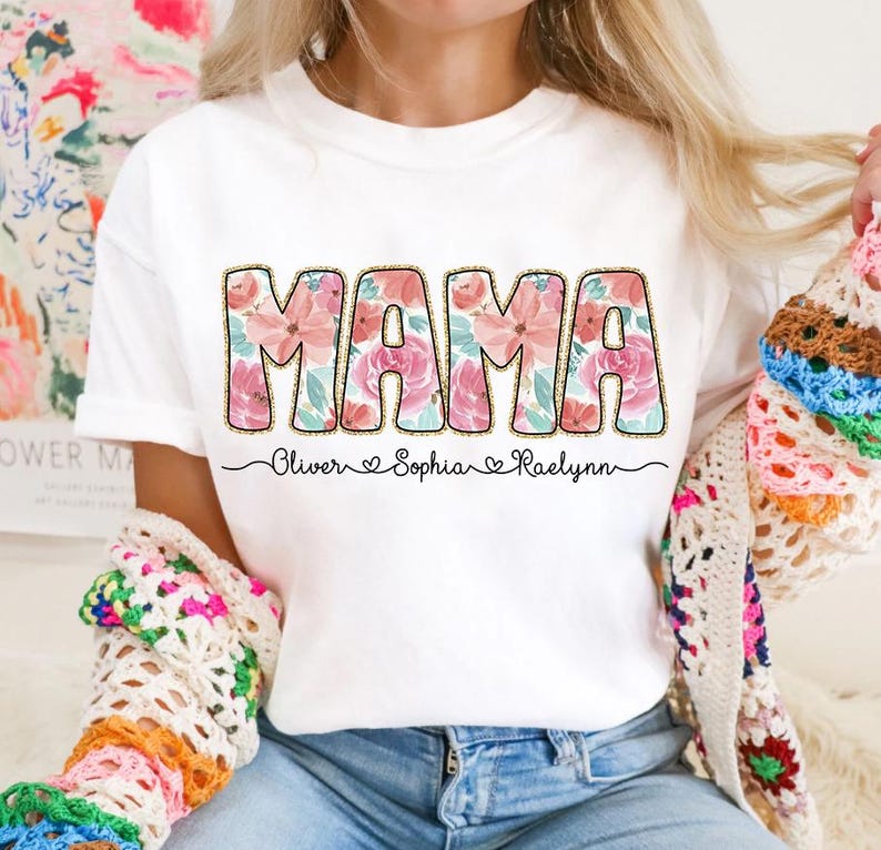 May include: White t-shirt with the word "MAMA" in pink and green floral letters. The names "Oliver", "Sophia", and "Raelynn" are written below the word "MAMA" with hearts between each name.