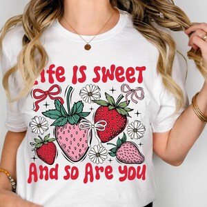 May include: White t-shirt with a red graphic that says "Life is sweet and so are you". The graphic features three strawberries with pink and red accents, green leaves, and white flowers.