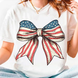 Peut inclure: T-shirt blanc avec un n&oelig;ud rouge, blanc et bleu imprim&eacute; sur le devant. Le n&oelig;ud est dessin&eacute; dans un style vintage et ressemble au drapeau am&eacute;ricain.