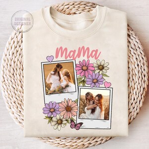 May include: Cream-colored sweatshirt with the word "Mama" in pink script. The design includes two polaroid-style photo frames with images of a mother and child, surrounded by colorful flowers and a butterfly.