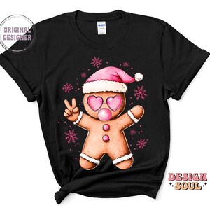 May include: Black t-shirt with a gingerbread man wearing a pink Santa hat and pink heart-shaped sunglasses. The gingerbread man is holding up two fingers in a peace sign. The design is surrounded by snowflakes.