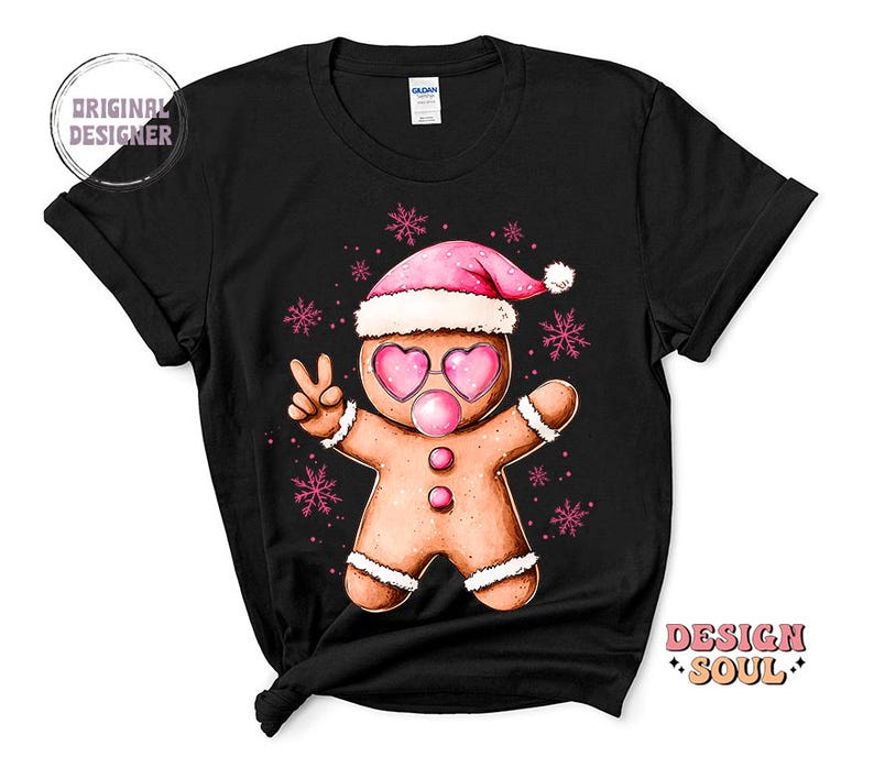 May include: Black t-shirt with a gingerbread man wearing a pink Santa hat and pink heart-shaped sunglasses. The gingerbread man is holding up two fingers in a peace sign. The design is surrounded by snowflakes.