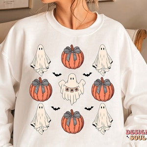 May include: White sweatshirt featuring a Halloween-themed design. The design includes orange pumpkins with gray bows, friendly ghosts, and black bats. One ghost holds a banner that spells "BOO". The words "DESIGN SOUL" are printed in pink.