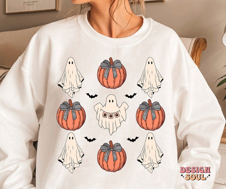 May include: White sweatshirt featuring a Halloween-themed design. The design includes orange pumpkins with gray bows, friendly ghosts, and black bats. One ghost holds a banner that spells "BOO". The words "DESIGN SOUL" are printed in pink.