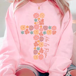 May include: A pink sweatshirt with a floral cross design and the text "He is Risen".