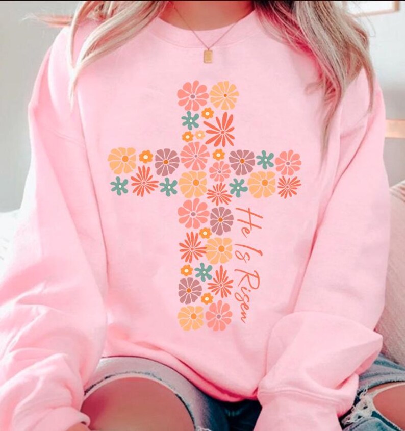 May include: A pink sweatshirt with a floral cross design and the text "He is Risen".