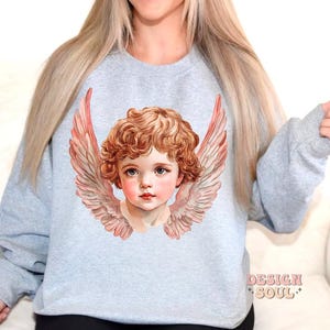 Op de afbeelding: Een lichtgrijs sweatshirt met een afbeelding van een cherubijn met vleugels. De cherubijn heeft blond haar en blauwe ogen. De vleugels zijn roze en wit.