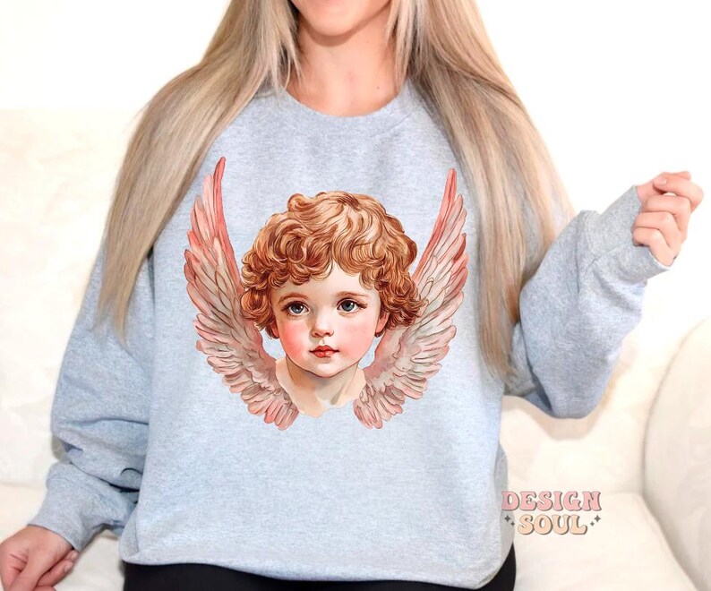 Op de afbeelding: Een lichtgrijs sweatshirt met een afbeelding van een cherubijn met vleugels. De cherubijn heeft blond haar en blauwe ogen. De vleugels zijn roze en wit.