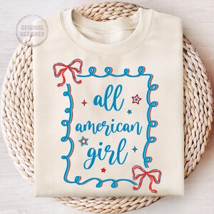 Peut inclure: Un sweat-shirt cr&egrave;me avec un motif rouge, blanc et bleu. Le motif pr&eacute;sente un cadre avec un n&oelig;ud en haut et en bas, et le texte "all american girl" &agrave; l'int&eacute;rieur du cadre.