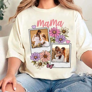 May include: Cream-colored t-shirt with the word "Mama" in pink script, decorated with floral and butterfly graphics. Two polaroid-style photo frames feature images of a mother and child. The shirt is a casual, comfortable style.