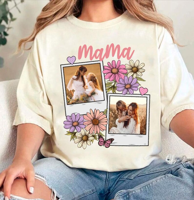 May include: Cream-colored t-shirt with the word "Mama" in pink script, decorated with floral and butterfly graphics. Two polaroid-style photo frames feature images of a mother and child. The shirt is a casual, comfortable style.