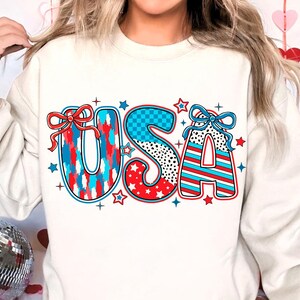 May include: White sweatshirt with a red, white, and blue "USA" graphic. The letters are decorated with stars, stripes, and checkered patterns.