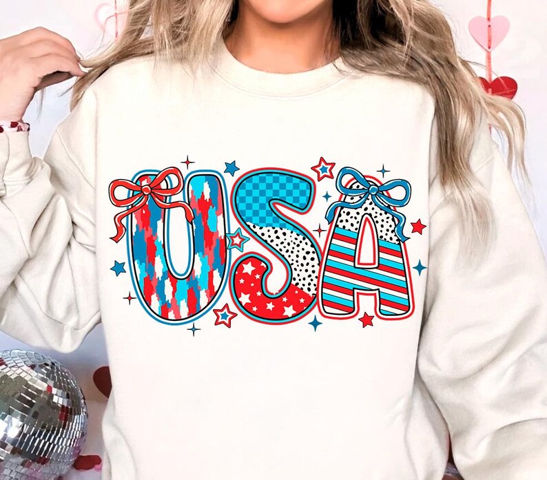 May include: White sweatshirt with a red, white, and blue "USA" graphic. The letters are decorated with stars, stripes, and checkered patterns.