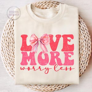 May include: A cream-colored sweatshirt with a pink bow and the text "LOVE MORE worry less" in a pink distressed font.