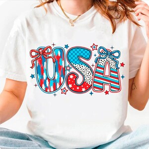 May include: White t-shirt with a red, white, and blue "USA" graphic. The letters are decorated with stars, stripes, and checkered patterns.