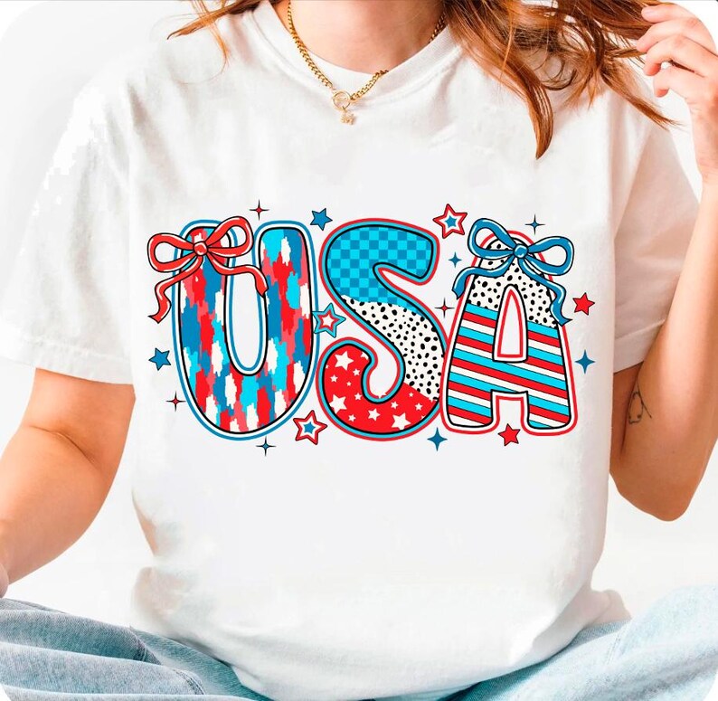 May include: White t-shirt with a red, white, and blue "USA" graphic. The letters are decorated with stars, stripes, and checkered patterns.