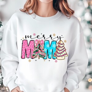Peut inclure: Sweat-shirt blanc avec un motif de No&euml;l. Le texte "merry" est au-dessus du mot "MAMA" qui est d&eacute;cor&eacute; de sapins de No&euml;l roses, bleus et verts.