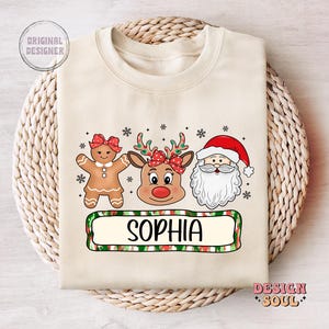 May include: A beige Christmas sweatshirt with a gingerbread person, reindeer, and Santa Claus illustration. The text "Sophia" is printed on a green and red striped banner.