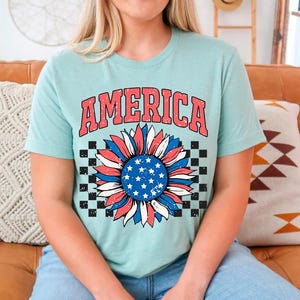 May include: A light blue t-shirt with a red, white, and blue sunflower graphic and the word "AMERICA" in red letters. The sunflower has a checkered background.