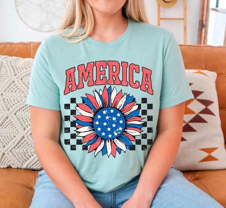 May include: A light blue t-shirt with a red, white, and blue sunflower graphic and the word "AMERICA" in red letters. The sunflower has a checkered background.