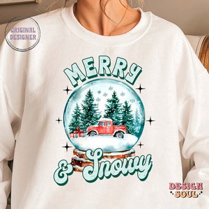 May include: White sweatshirt with a snow globe design featuring a red truck, green trees, and snowflakes. The text "Merry & Snowy" is written in green and white below the snow globe.