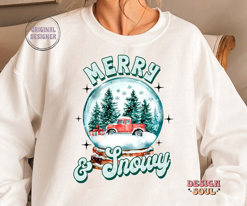 May include: White sweatshirt with a snow globe design featuring a red truck, green trees, and snowflakes. The text "Merry & Snowy" is written in green and white below the snow globe.