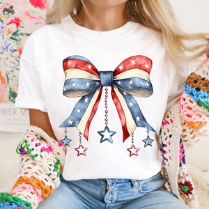 May include: A white t-shirt with a red, white, and blue bow with stars printed on it. The bow has strings with stars hanging from it.