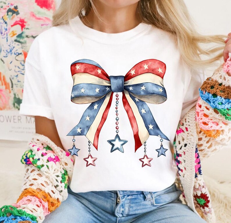 May include: A white t-shirt with a red, white, and blue bow with stars printed on it. The bow has strings with stars hanging from it.