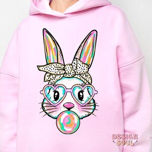 May include: Pink hoodie featuring a cartoon bunny wearing heart-shaped sunglasses and a headband, blowing a bubblegum bubble.