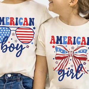 May include: Two white t-shirts with red, white, and blue American flag designs. The shirt on the left says "AMERICAN boy" with sunglasses featuring the American flag. The shirt on the right says "AMERICAN girl" with a red, white, and blue bow.