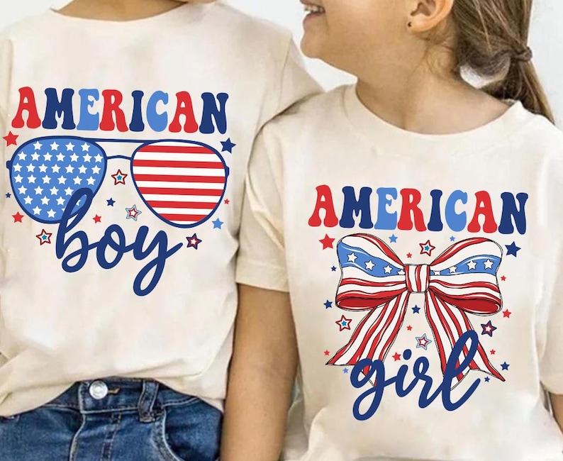 May include: Two white t-shirts with red, white, and blue American flag designs. The shirt on the left says "AMERICAN boy" with sunglasses featuring the American flag. The shirt on the right says "AMERICAN girl" with a red, white, and blue bow.