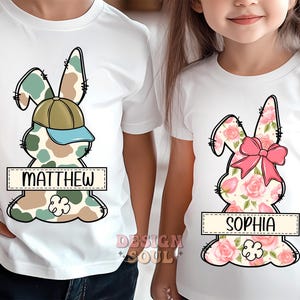 May include: Two white t-shirts with a bunny rabbit design. One bunny is camouflage with a blue baseball cap and the name "MATTHEW" printed below. The other bunny is pink and floral with the name "SOPHIA" printed below.