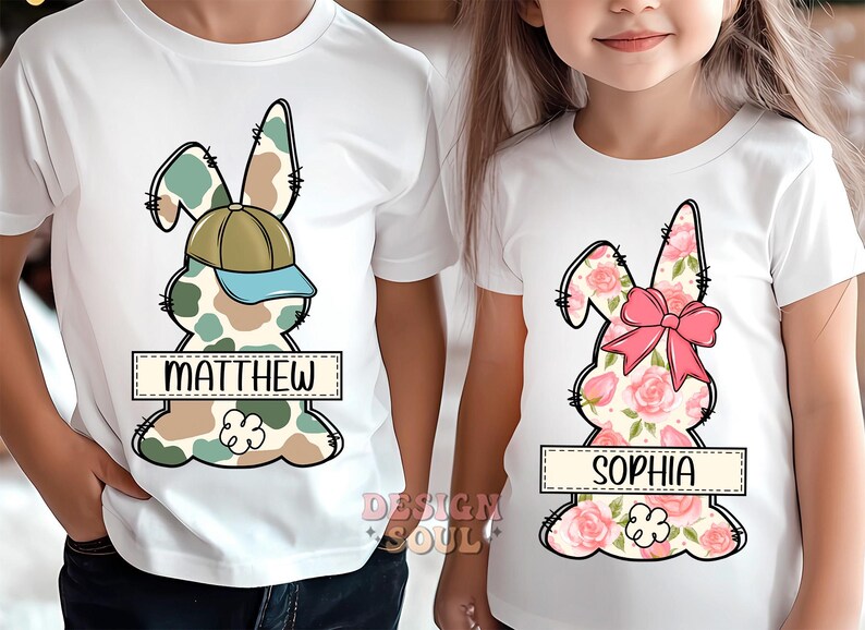 May include: Two white t-shirts with a bunny rabbit design. One bunny is camouflage with a blue baseball cap and the name "MATTHEW" printed below. The other bunny is pink and floral with the name "SOPHIA" printed below.