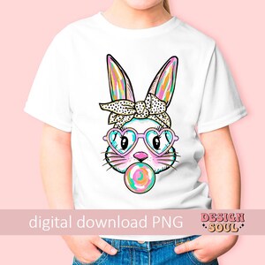May include: A white t-shirt featuring a colourful graphic of a bunny wearing pink heart-shaped sunglasses and a polka dot headband. The bunny is holding a pink and blue lollipop in its mouth. The text "digital download PNG" is at the bottom of the shirt.