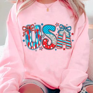 May include: Pink sweatshirt with a graphic of the word "USA" in red, white, and blue with stars and checkered patterns.