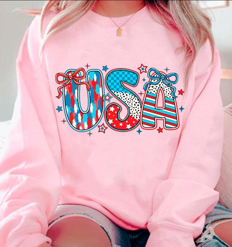 May include: Pink sweatshirt with a graphic of the word "USA" in red, white, and blue with stars and checkered patterns.
