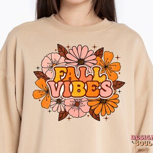 May include: A beige sweatshirt with a retro-style graphic design featuring the text "FALL VIBES" surrounded by orange, pink, and brown flowers. The design has a vintage, distressed look.