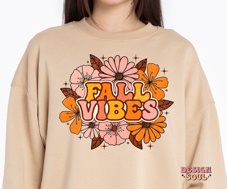 May include: A beige sweatshirt with a retro-style graphic design featuring the text "FALL VIBES" surrounded by orange, pink, and brown flowers. The design has a vintage, distressed look.