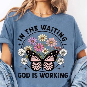 May include: A blue t-shirt with a graphic of a butterfly and flowers in a circle. The text "In the waiting, God is working" is printed on the shirt.