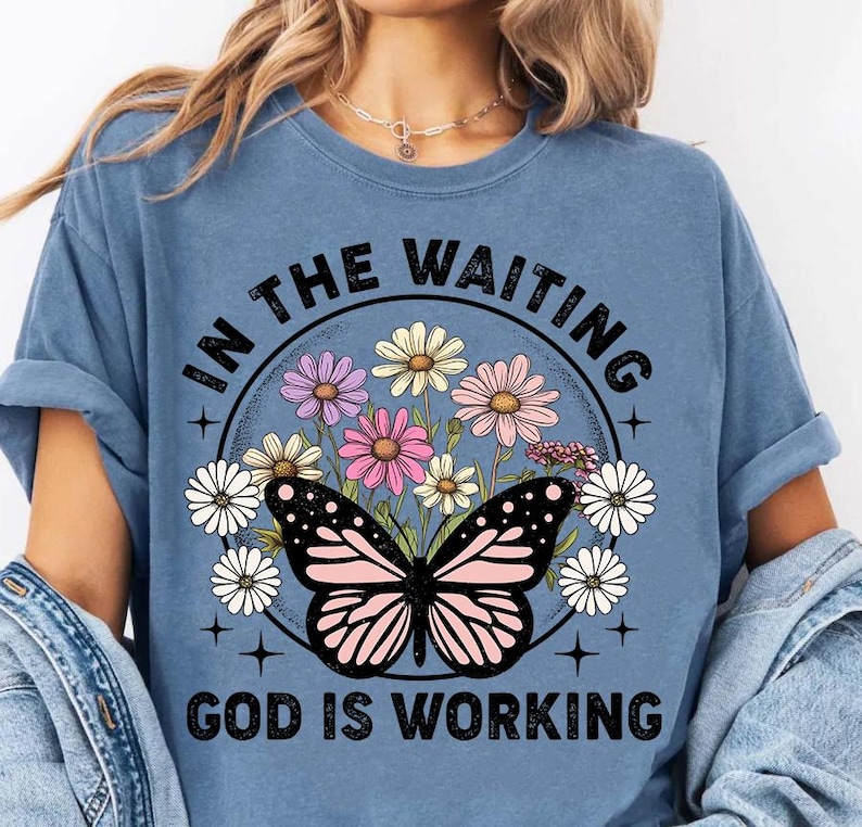 May include: A blue t-shirt with a graphic of a butterfly and flowers in a circle. The text "In the waiting, God is working" is printed on the shirt.