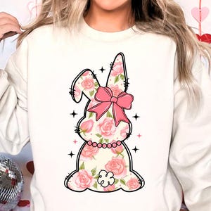 May include: White sweatshirt with a pink floral bunny graphic. The bunny has a pink bow and is surrounded by stars.
