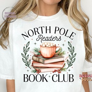 May include: White t-shirt with a Christmas-themed design. The design features a mug of hot chocolate with marshmallows, a stack of books, and greenery. The text "North Pole Readers Book Club" is printed on the shirt.