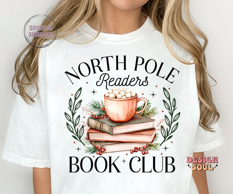 May include: White t-shirt with a Christmas-themed design. The design features a mug of hot chocolate with marshmallows, a stack of books, and greenery. The text "North Pole Readers Book Club" is printed on the shirt.
