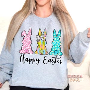 May include: A light gray sweatshirt with a graphic of three Easter bunnies. The bunnies are pink, blue, and green with floral patterns. The text "Happy Easter" is printed below the bunnies.