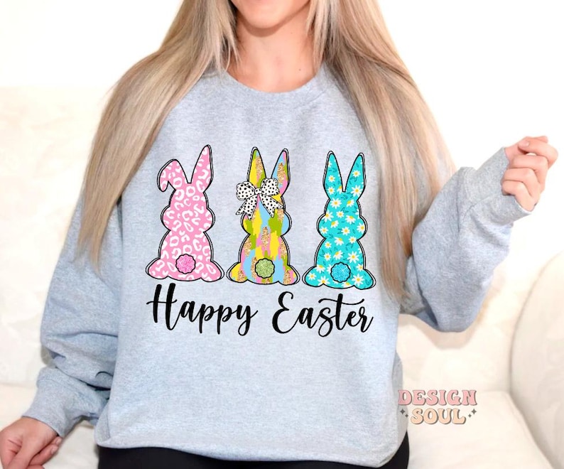 May include: A light gray sweatshirt with a graphic of three Easter bunnies. The bunnies are pink, blue, and green with floral patterns. The text "Happy Easter" is printed below the bunnies.