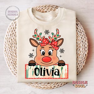 May include: A beige sweatshirt with a cartoon reindeer wearing a red bow. The reindeer is looking at the viewer and has a red nose. The text "Olivia" is written on a colorful banner below the reindeer.